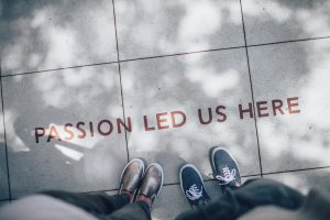 A photo of the pavement taken from above. Two people’s feet are visible in the shot. In front of their feet is text reading ‘passion led us here’. 