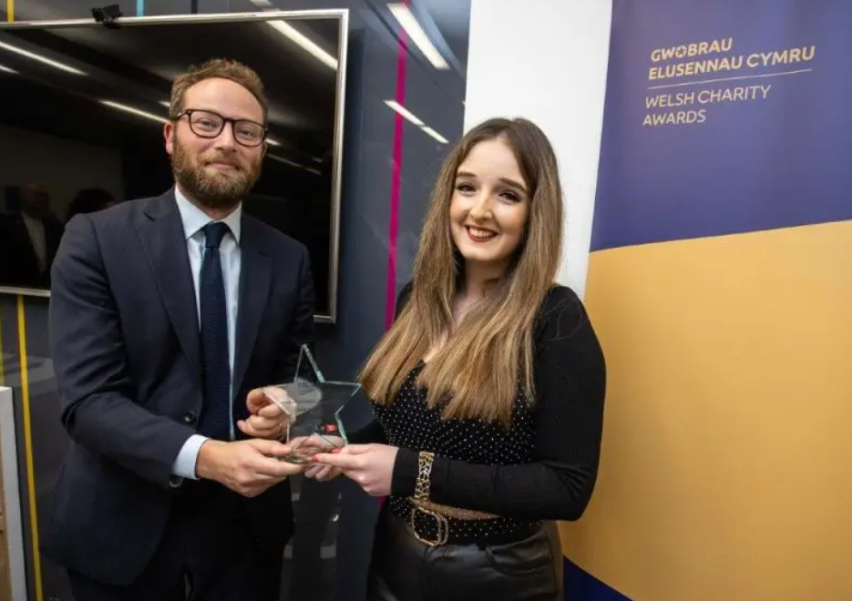 Rachel is smiling as she stands in front of the Welsh Charity Awards banner, being presented with her award.