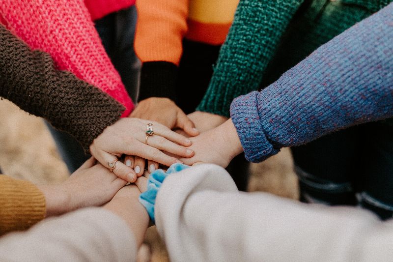 A group of people wearing colorful sweaters place their hands together in a show of unity.