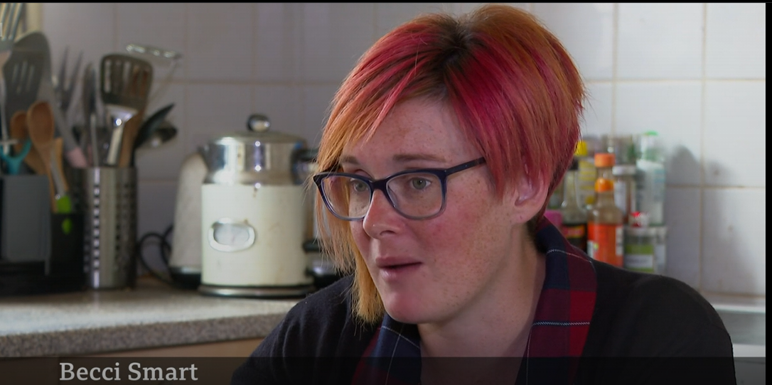 Becci is a white woman with short red and orange dyed hair. She is sat in her kitchen speaking to a reporter who is off camera.